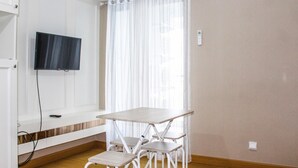 Una habitación con una televisión montada en la pared, una mesa blanca con sillas y una ventana con cortinas translúcidas.