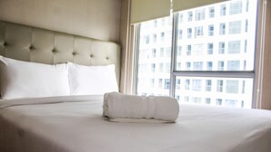 Una cama impecablemente arreglada con una toalla enrollada en una habitación de hotel con un ventanal grande y un cabecero capitonado.
