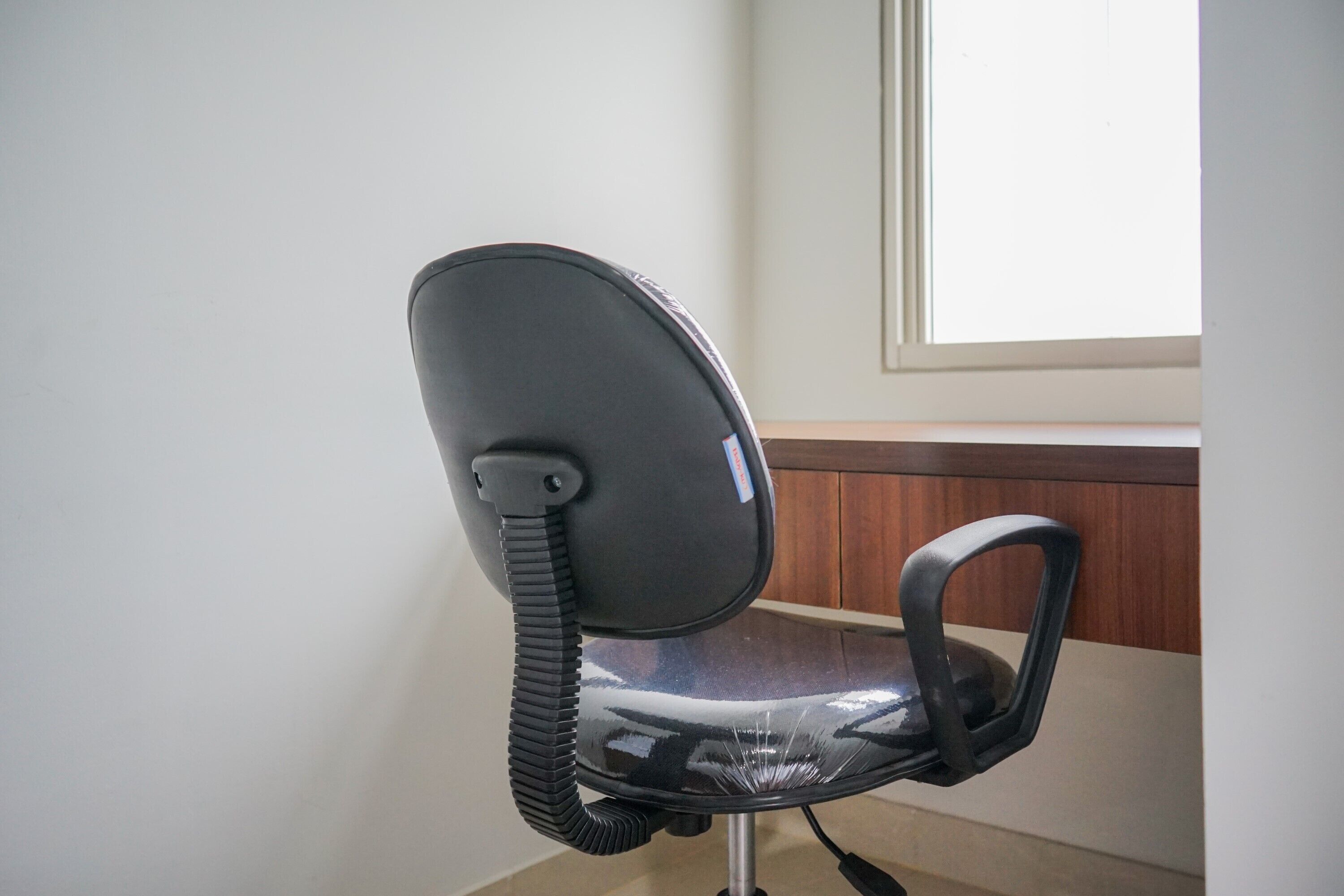 A black office chair with a headrest and armrest, positioned in a room with a wooden panel and a window.