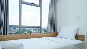 A neatly made bed with a white towel, a wooden headboard, and a view of a cityscape through a large window with curtains.