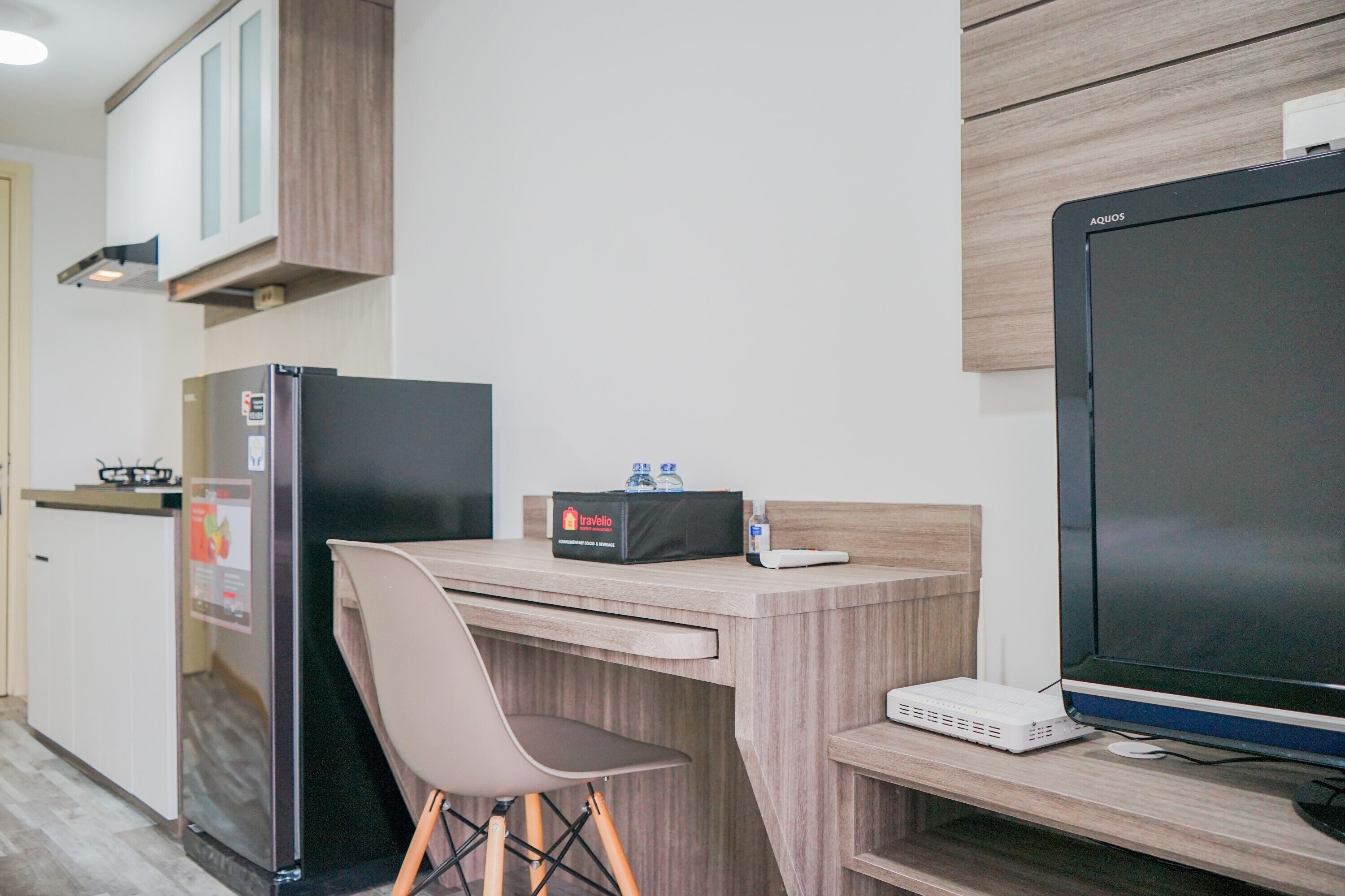 A kitchenette with a refrigerator, a travel bag, and a computer setup including a monitor and a router.