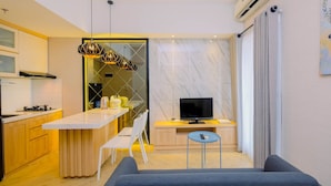 A modern kitchen with a white countertop, a dining area with a blue chair, a TV on a wooden stand, and a painting on the wall. - Relax 3BR Apartment at M-Town Residence (Gading Serpong)