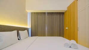 A hotel room with a neatly made bed, a headboard, a curtain, and a wall-mounted light fixture. - Relax 3BR Apartment at M-Town Residence (Gading Serpong)