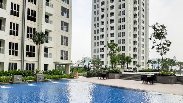 A swimming pool with a clear blue water surface, surrounded by a landscaped area with benches and a palm tree. - Brand New 1BR Apartment at M-Town Signature (Gading Serpong)