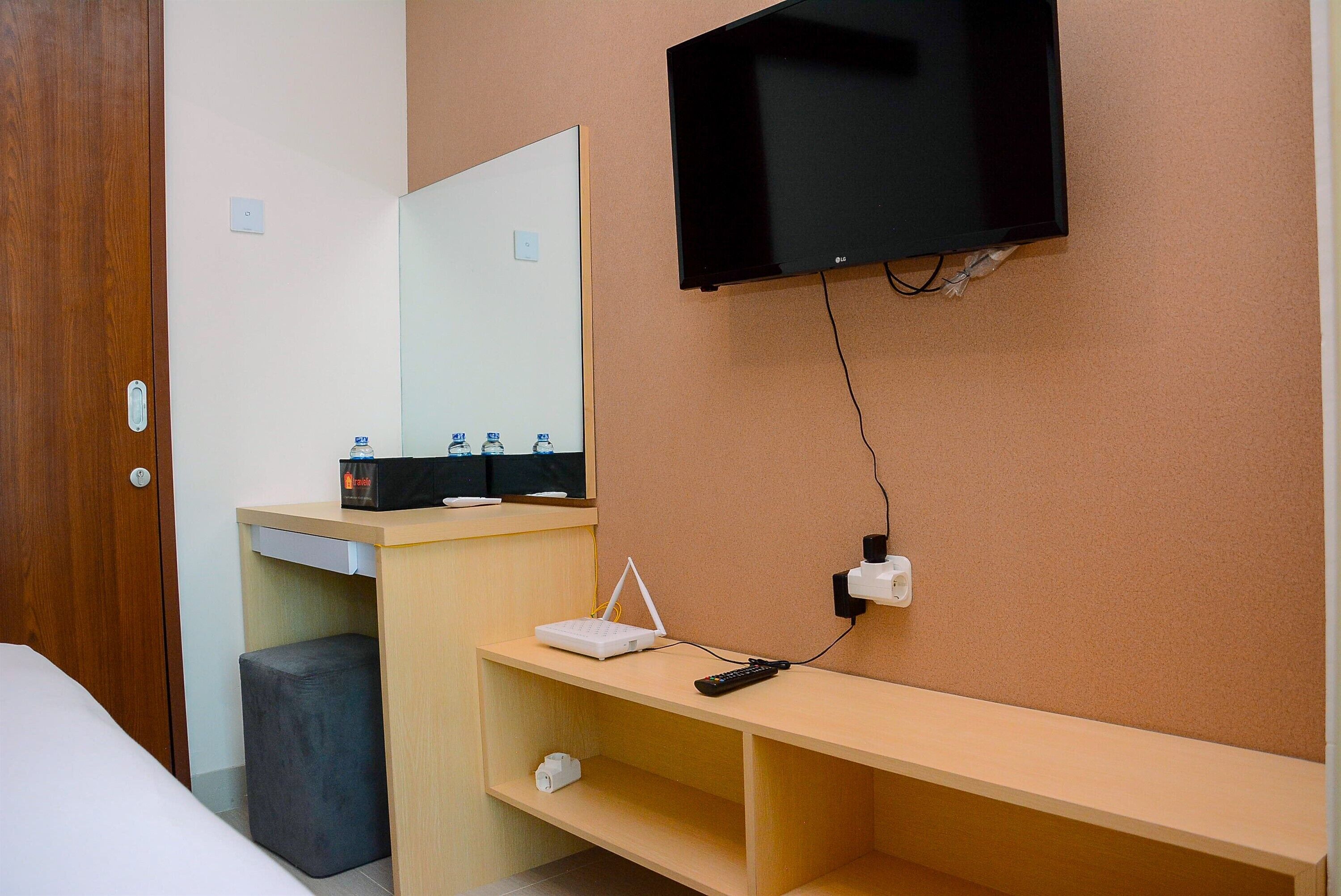 A hotel room with a bed, a desk with a computer and a TV, a mirror, and a small stool.