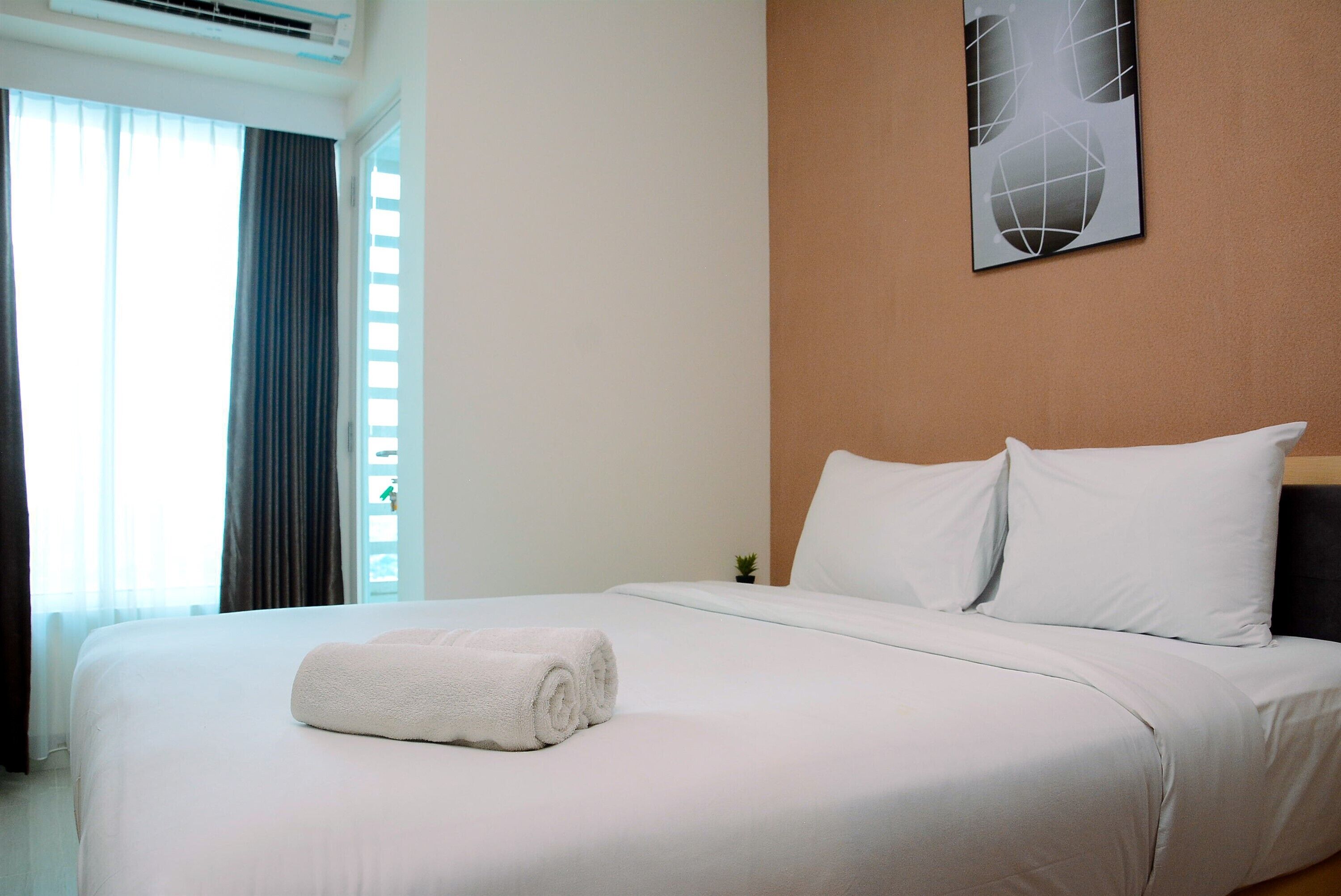 A neatly made bed with white linens and a rolled towel, a small potted plant, and a geometric wall art piece.