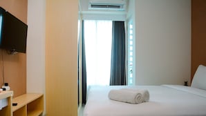 A hotel room with a bed, a TV mounted on the wall, a wooden shelf, and a window with curtains. - Minimalist and Homey 1BR Grand Kamala Lagoon Apartment (Bekasi)