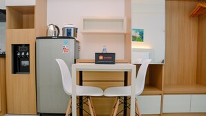 Eine kompakte Küchenzeile mit Kühlschrank, Kaffeemaschine und Wasserspender. Ein kleiner Tisch mit zwei weißen Stühlen und ein Schild für kostenlose Speisen und Getränke.