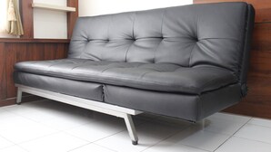 A grey leather sofa with a wooden frame, positioned against a white tiled floor and a wooden wall. - Relaxing 2BR Apartment at Buah Batu Park (Dayeuhkolot)
