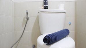 Casa de banho com sanita branca, mangueira de chuveiro e toalha enrolada no assento da sanita.