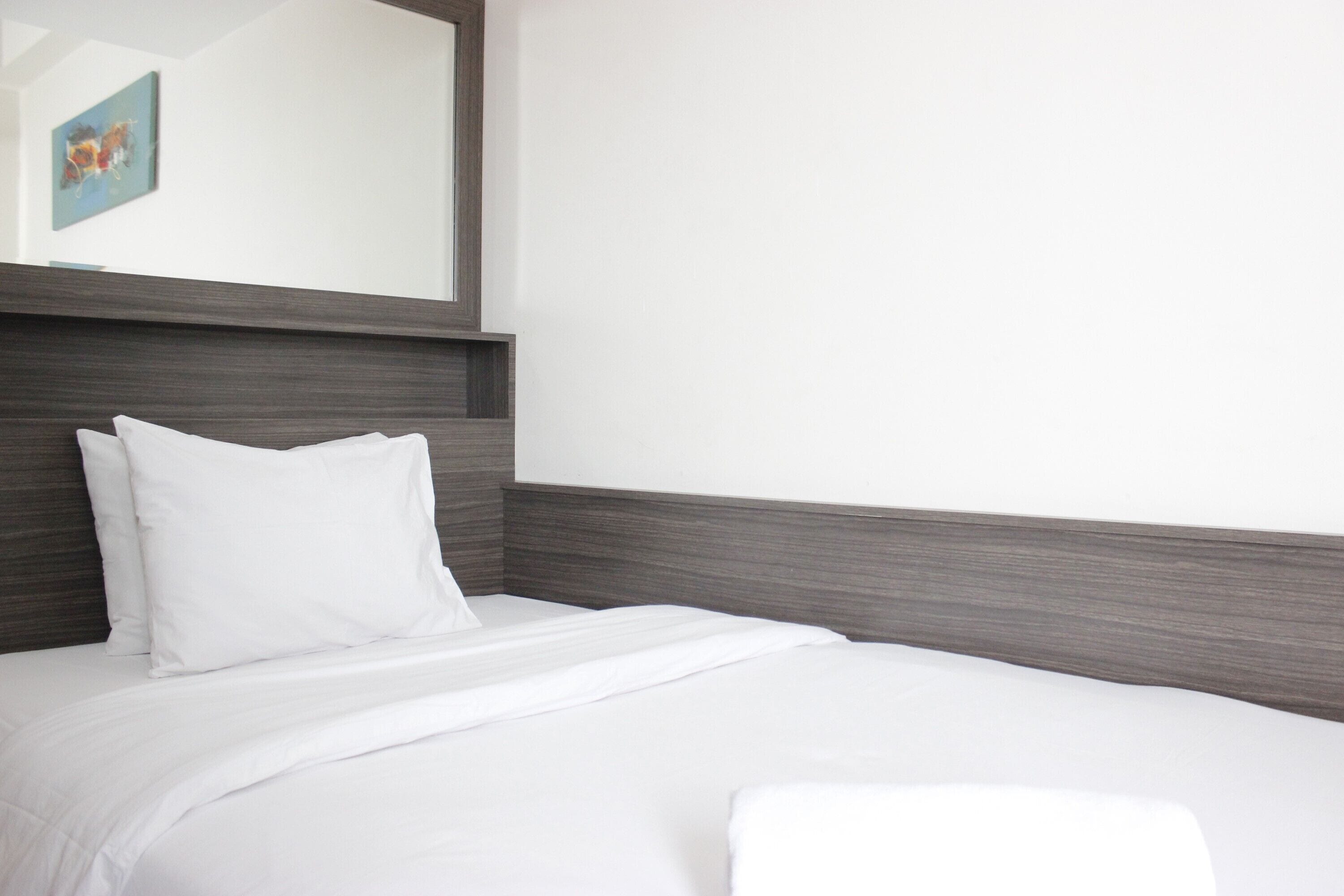 Una cama bien hecha con sábanas y almohadas blancas, un cabecero de madera oscura y un espejo grande en la pared.