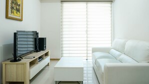 Un salon moderne avec un canapé blanc, un meuble TV en bois sur lequel sont posées deux télévisions à écran plat, une table basse blanche et une grande fenêtre équipée de stores.