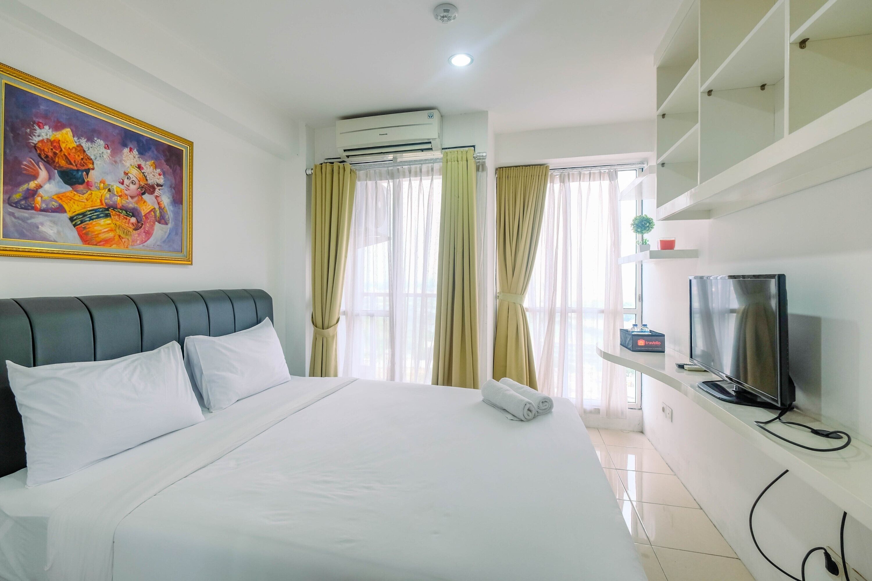 A hotel room with a bed, air conditioning unit, curtains, a television, and a painting on the wall.