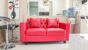 A red leather sofa with matching cushions, a black bar stool, and a kitchen area with a water dispenser and a coffee maker. - Modern and Relax 2BR @ Bassura City Apartment (Jakarta)