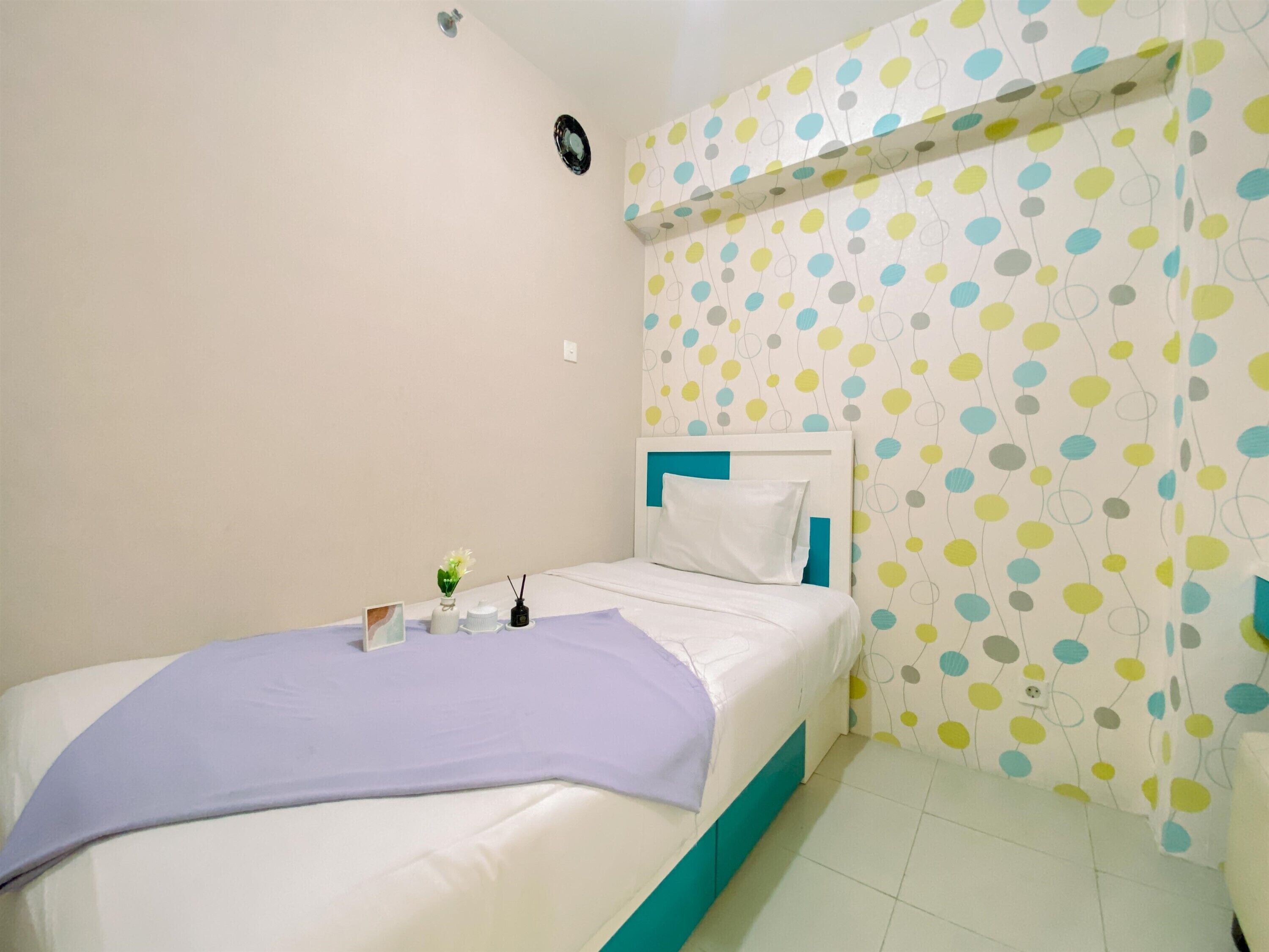 A bedroom with a bed, a bedside table with a vase and a book, and a wall with a polka dot pattern.