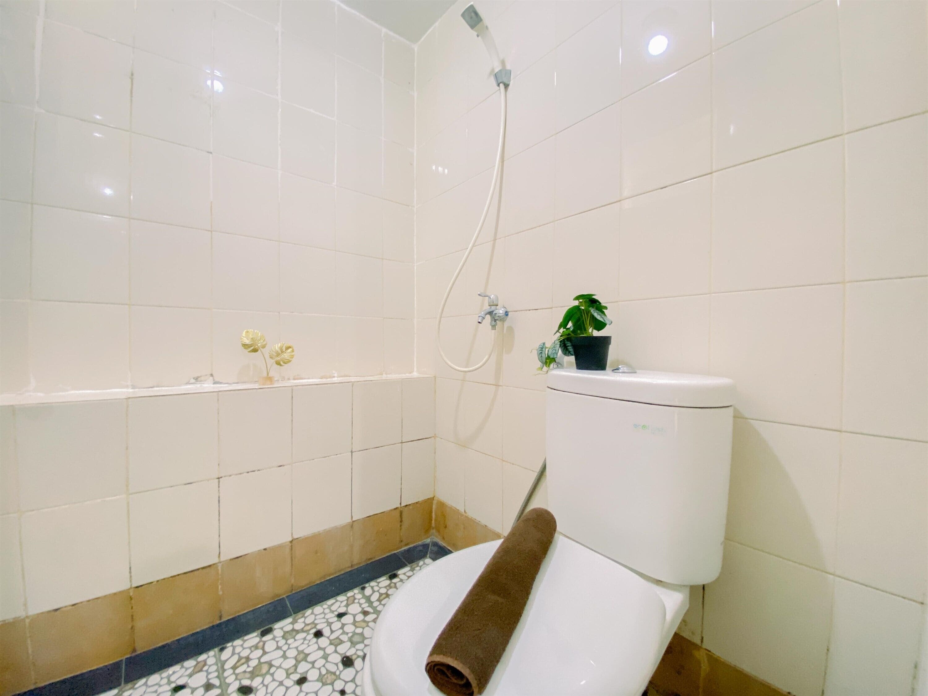 A bathroom with white tiles, a shower, a toilet, and a small shelf with a plant and flowers.