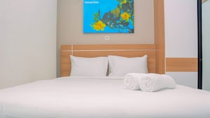 Een net opgemaakt bed met witte lakens en twee opgerolde handdoeken, een houten hoofdbord en een aan de muur hangend schilderij van gele bloemen.