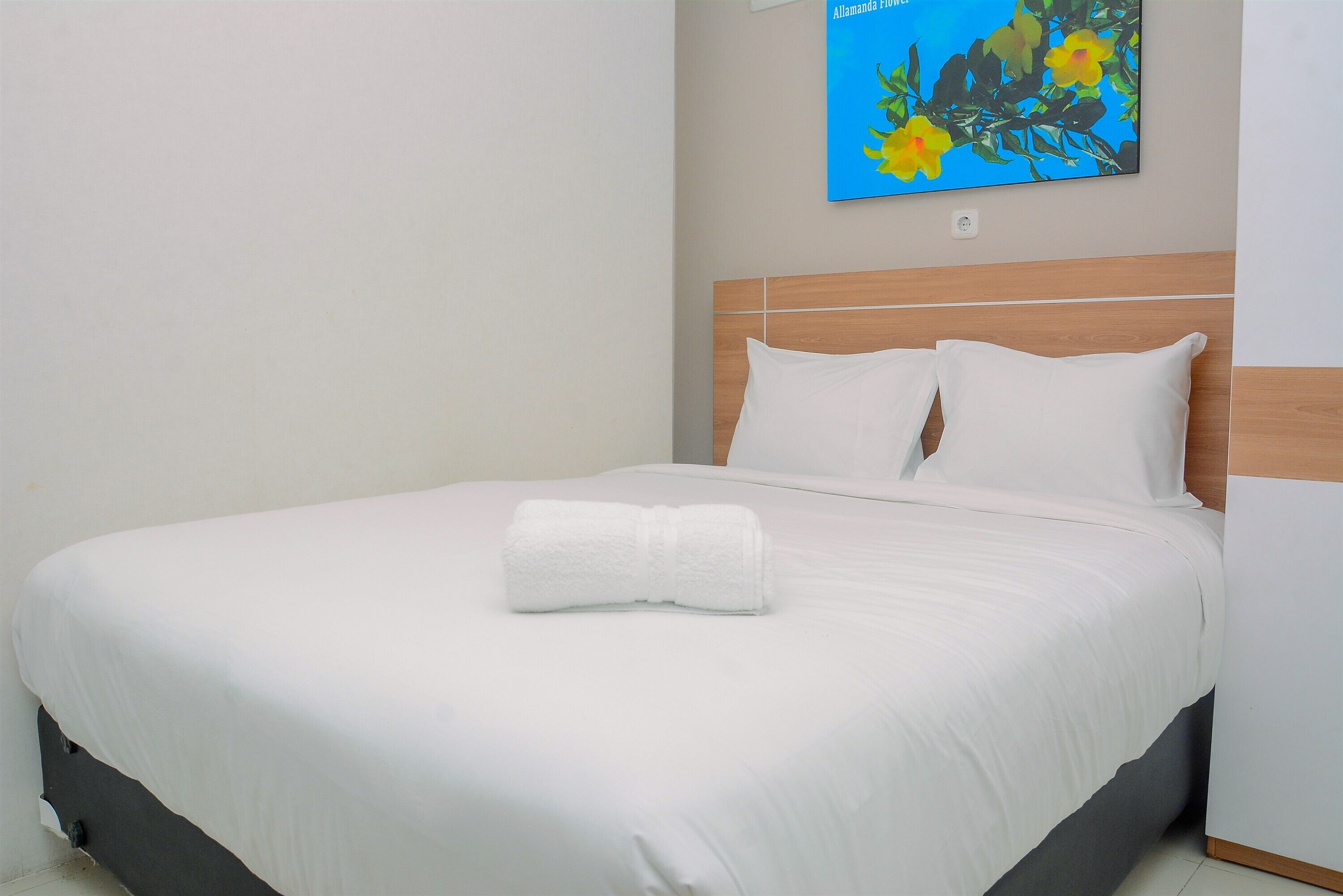 A neatly made bed with white linens and a rolled towel, a wooden headboard, and a wall-mounted painting of flowers.