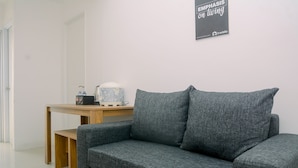 A modern room with a grey sofa, a wooden table with a kettle and water bottles, and a wall-mounted sign. - 1BR Fully Furnished Bassura City Apartment (Jakarta)