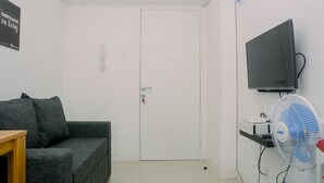 A room with a grey sofa, a white door, a mounted TV, and a standing fan. - 1BR Fully Furnished Bassura City Apartment (Jakarta)