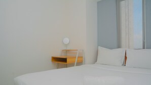 A neatly made bed with white linens and a white towel, a bedside table with a lamp, and a window with curtains. - Best Deal 3BR Apartment Bassura City near Shopping Center (Jakarta)
