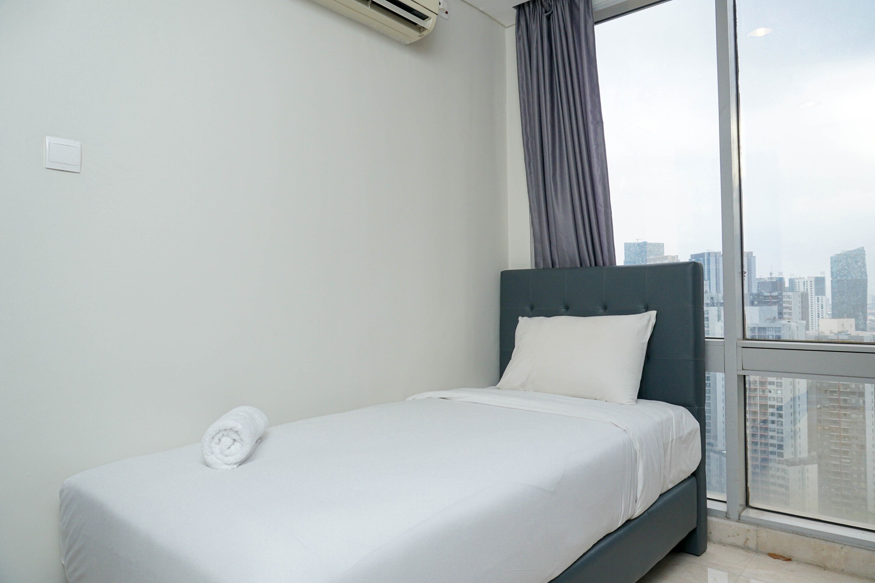 Ein Einzelbett mit weißer Matratze und Kissen, ein grauer Kopfteil und ein Fenster mit Blick auf die Stadt.