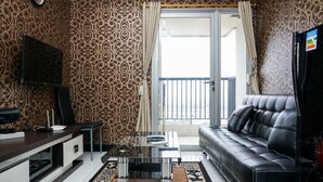 A modern living room with patterned wallpaper, a glass coffee table, a black leather sofa, a flat-screen TV, and a sliding glass door leading to a balcony. - Amazing 1BR Apartment at The Wave @ Rasuna Epicentrum (Jakarta)