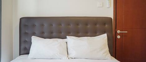 Ein ordentlich bezogenes Bett mit einer gebügelten Kopfstütze und zwei Kissen, eine Holztür rechts und ein an der Wand befestigtes Thermostat.