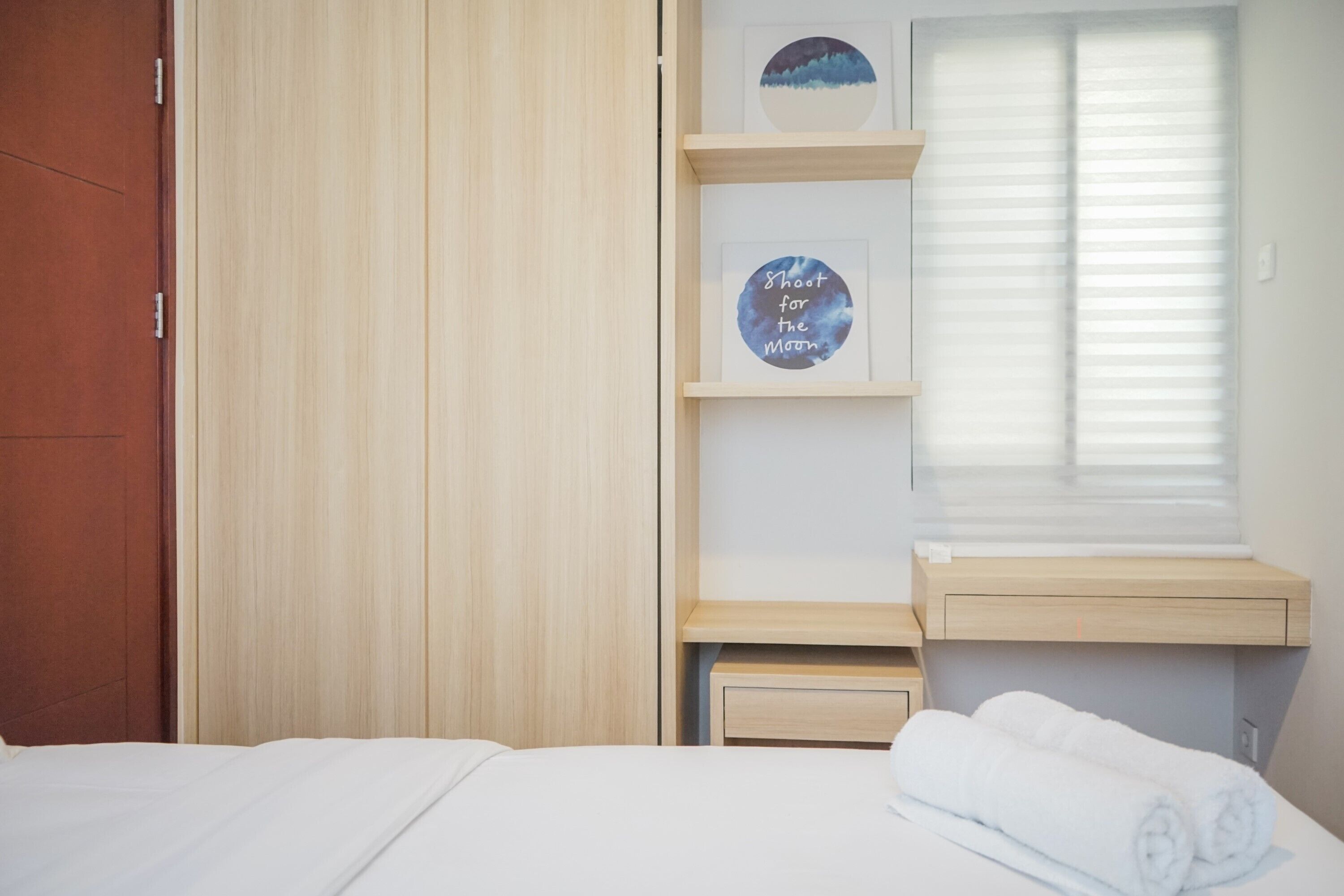 Ein Hotelzimmer mit Bett, Holzschrank, einer Ablage mit gerahmten Bildern und einem Fenster mit Jalousien.