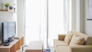 Ein modernes Wohnzimmer mit einem beigefarbenen Sofa, einem Flachbildfernseher auf einem Holzboden, einem kleinen Tisch und einem Fenster mit transparenten Vorhängen.