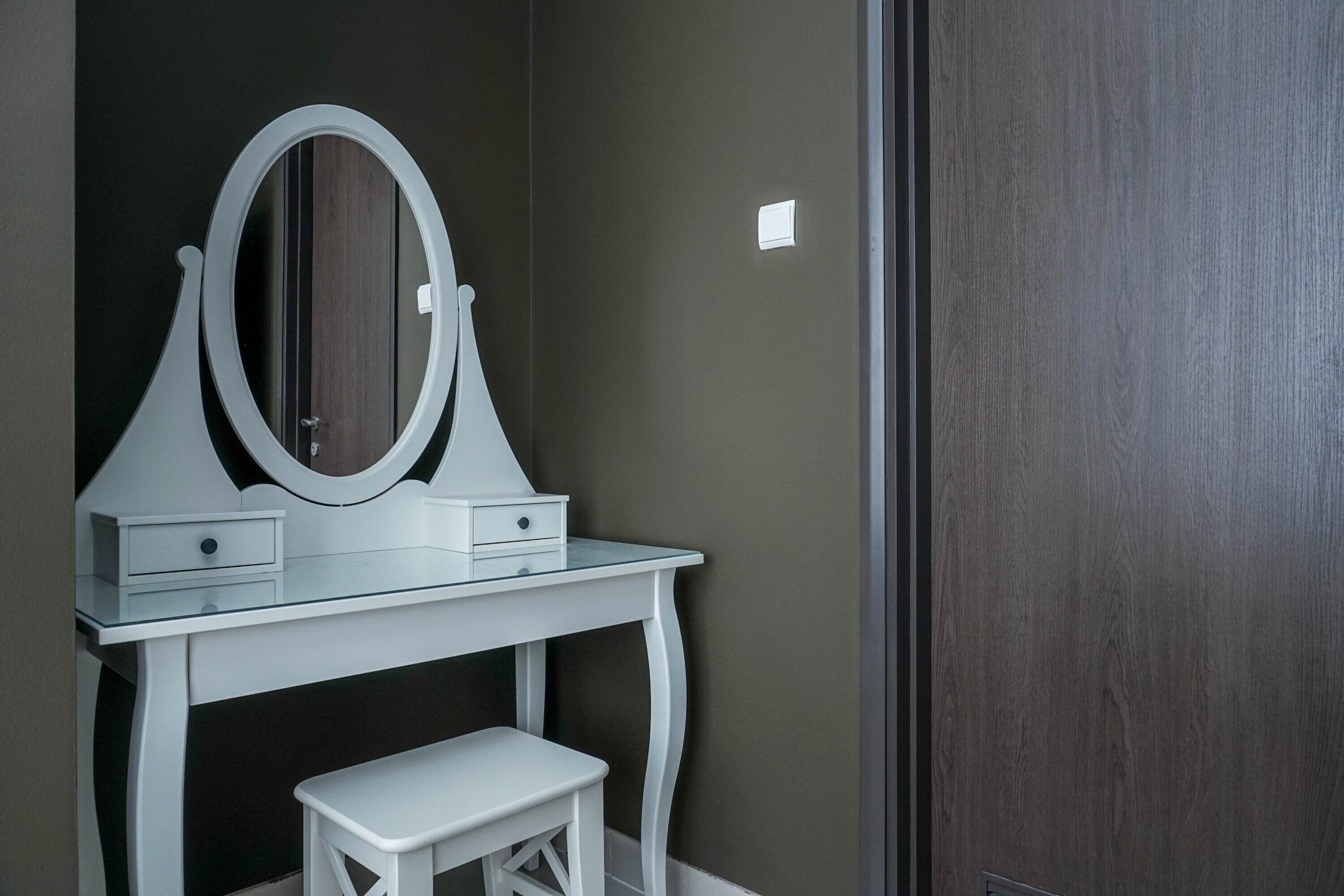A white vanity with a mirror and two drawers, a small white stool, and a dark wooden door.