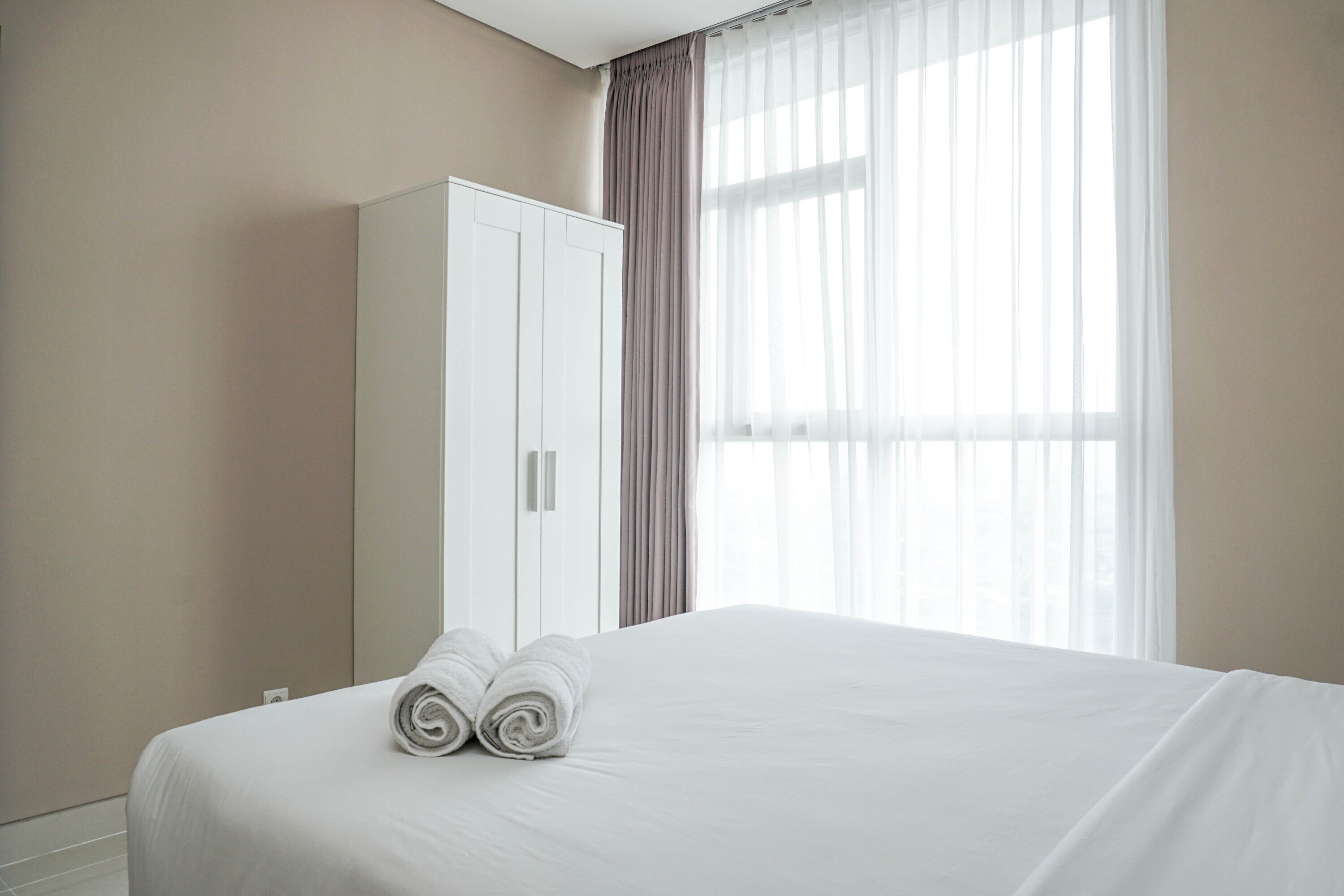 A hotel room with a neatly made bed, a wardrobe, and a large window with sheer curtains.