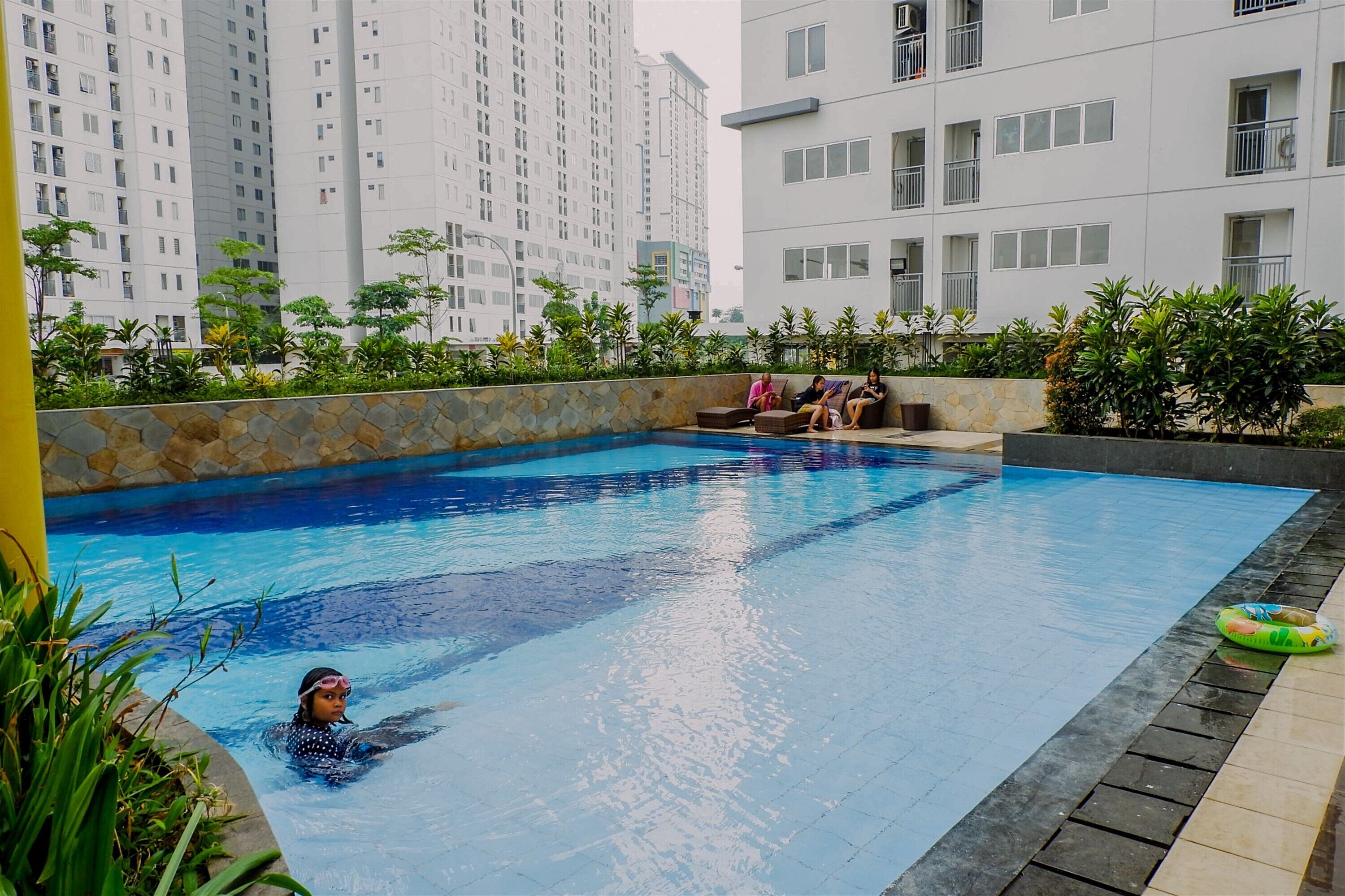 A swimming pool with a stone wall and plants, surrounded by high-rise buildings.