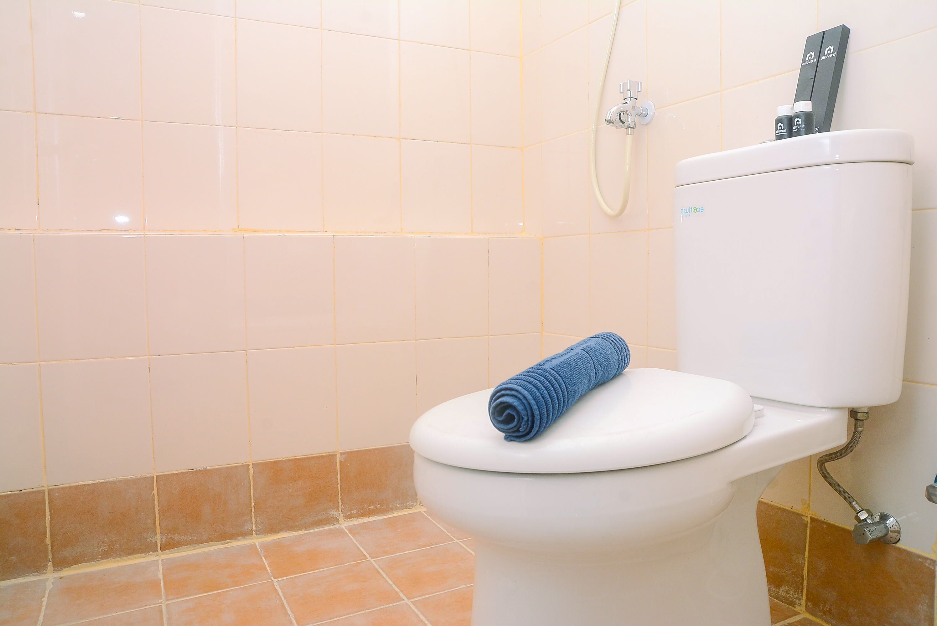 A toilet with a blue towel on the seat, a showerhead, and a toilet paper holder with a bottle on top.
