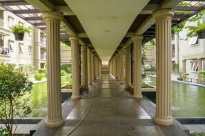 A corridor with columns, a water feature, and a view of a building with balconies. - Simply 1BR Grand Palace Kemayoran Apartment (Jakarta)