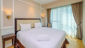 Una cama grande con sábanas blancas, un cabecero de madera y una toalla blanca sobre la cama. Hay dos mesitas de noche con lámparas, una ventana con cortinas traslúcidas y vistas a edificios.