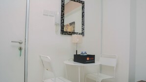 Una habitación pequeña con una puerta blanca, una silla blanca, una mesita con una lámpara, un espejo y una caja etiquetada 'Travello'.