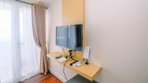 A hotel room with a mounted TV, a desk with a computer, a speaker, and a curtain. - Relaxing and Stylish Studio at Amethyst Tower Apartment (Jakarta)