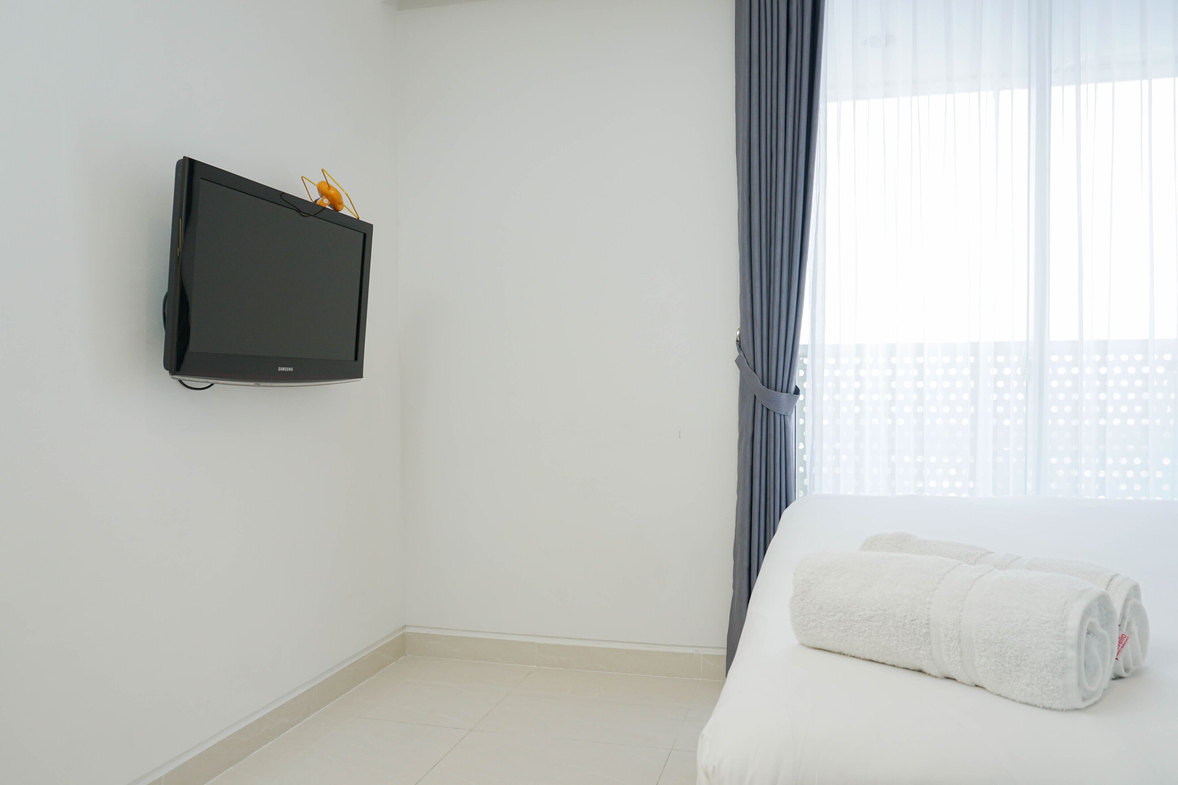 A hotel room with a flat-screen TV mounted on the wall, a neatly folded white towel on the bed, and a window with sheer curtains.
