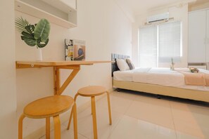 A bedroom with a bed, a bedside table, a chair, a potted plant, and a window with blinds. - Cozy Studio with Direct Access to Mall at Green Pramuka Apartment (Jakarta)
