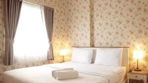 A neatly made bed with white linens and a rolled towel, a bedside lamp, and floral wallpaper. - Comfy & Strategic 2BR Gateway Pasteur Apartment near Pasteur Exit Toll (Cimahi)