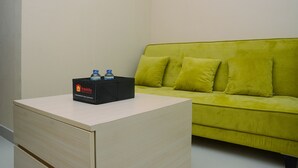 A small, light-colored desk with a black box labeled "Travelio" and two water bottles on top. A green sofa with three cushions is against the wall. - Fabulous 1BR Apartment at Atlanta Residences (Depok)