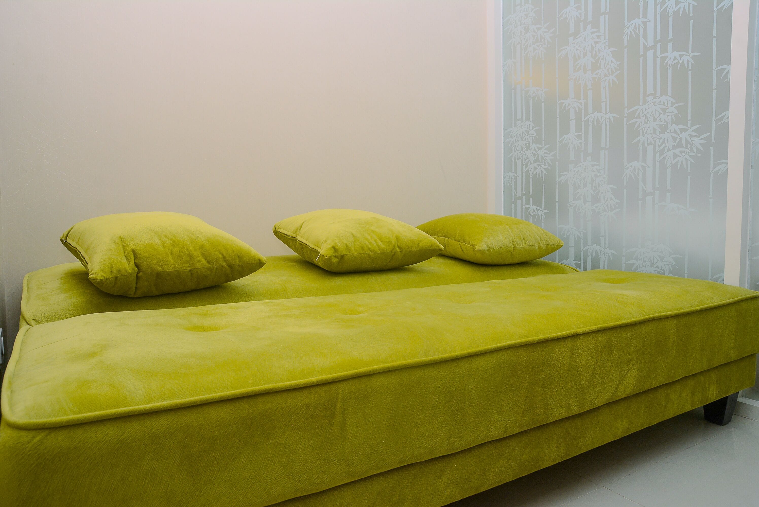 Un sofá amarillo brillante con tres cojines frente a una pared lisa y una persiana con estampado de bambú.