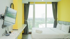 A hotel room with a bed, a desk with a computer, a TV, and a view of greenery through large windows. - Studio Apartment at Tree Park Apartment (South Tangerang)