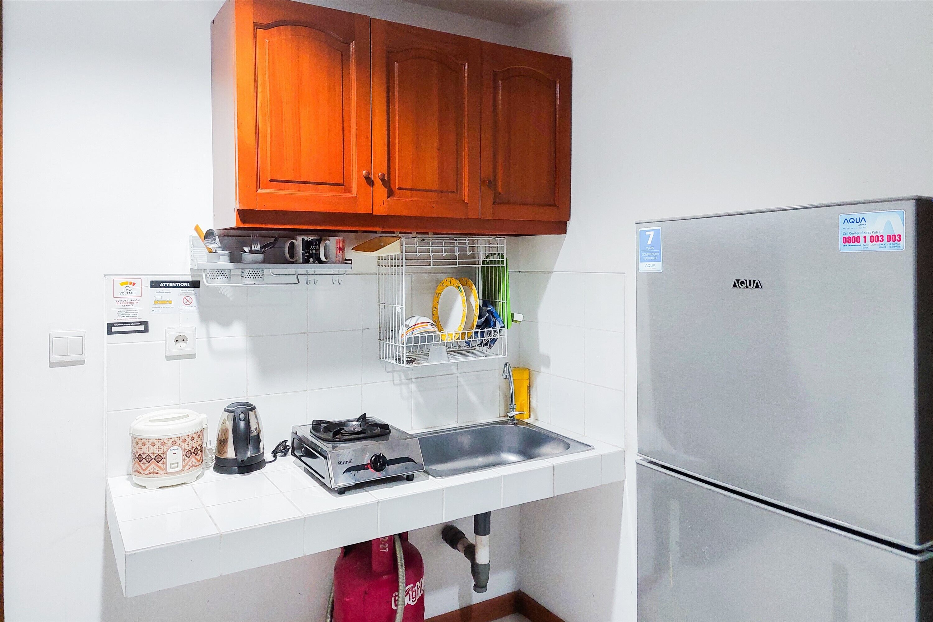 A compact kitchen with wooden cabinets, a stainless steel refrigerator, a sink, and a countertop with a coffee maker and a rice cooker.