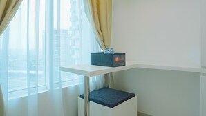 A hotel room with a white desk, a small seating area, and a view of buildings through sheer curtains. - New Furnished Studio Grand Kamala Lagoon Apartment (Bekasi)