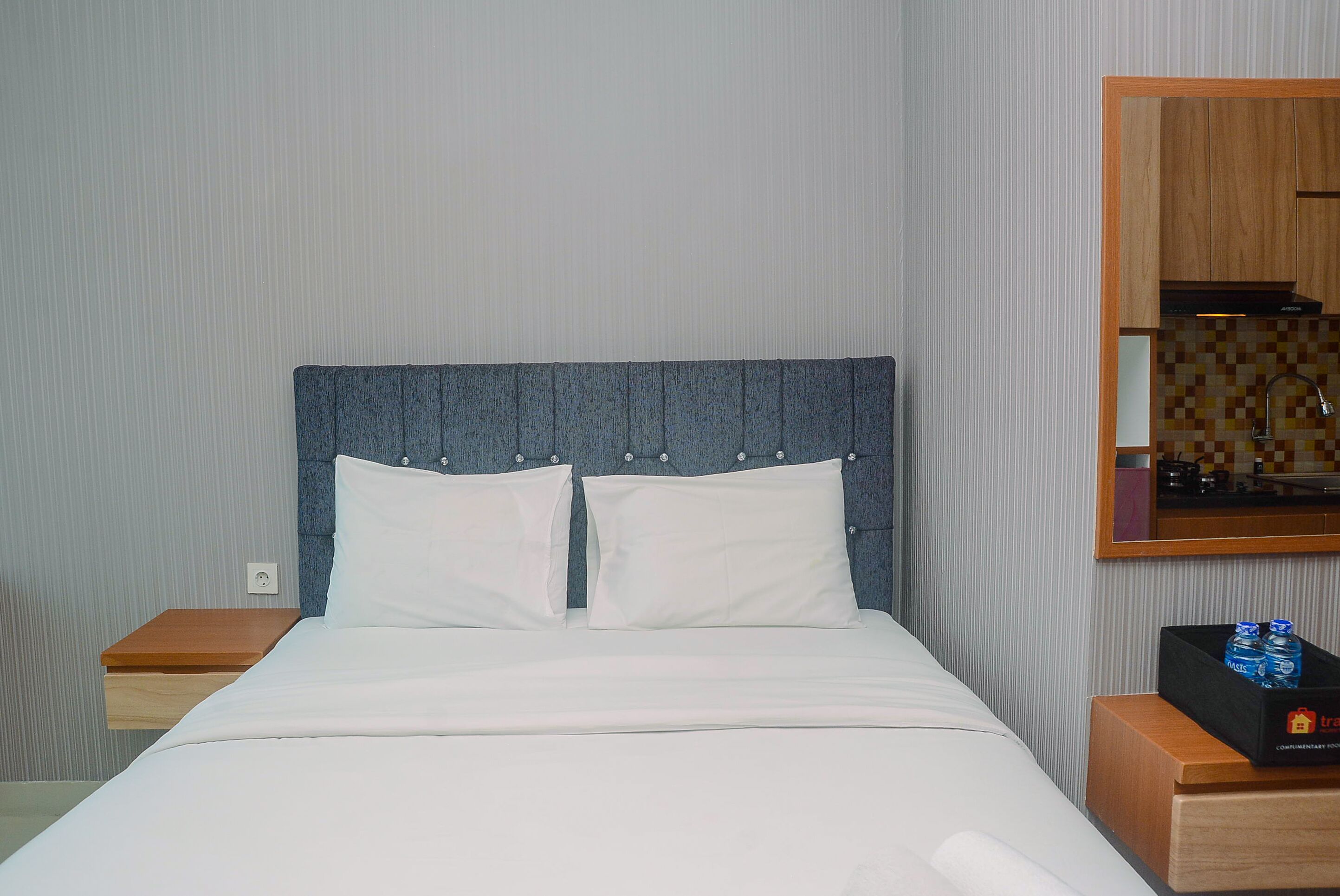 A neatly made bed with a headboard, a bedside table with a box of toiletries, and a wall-mounted mirror.