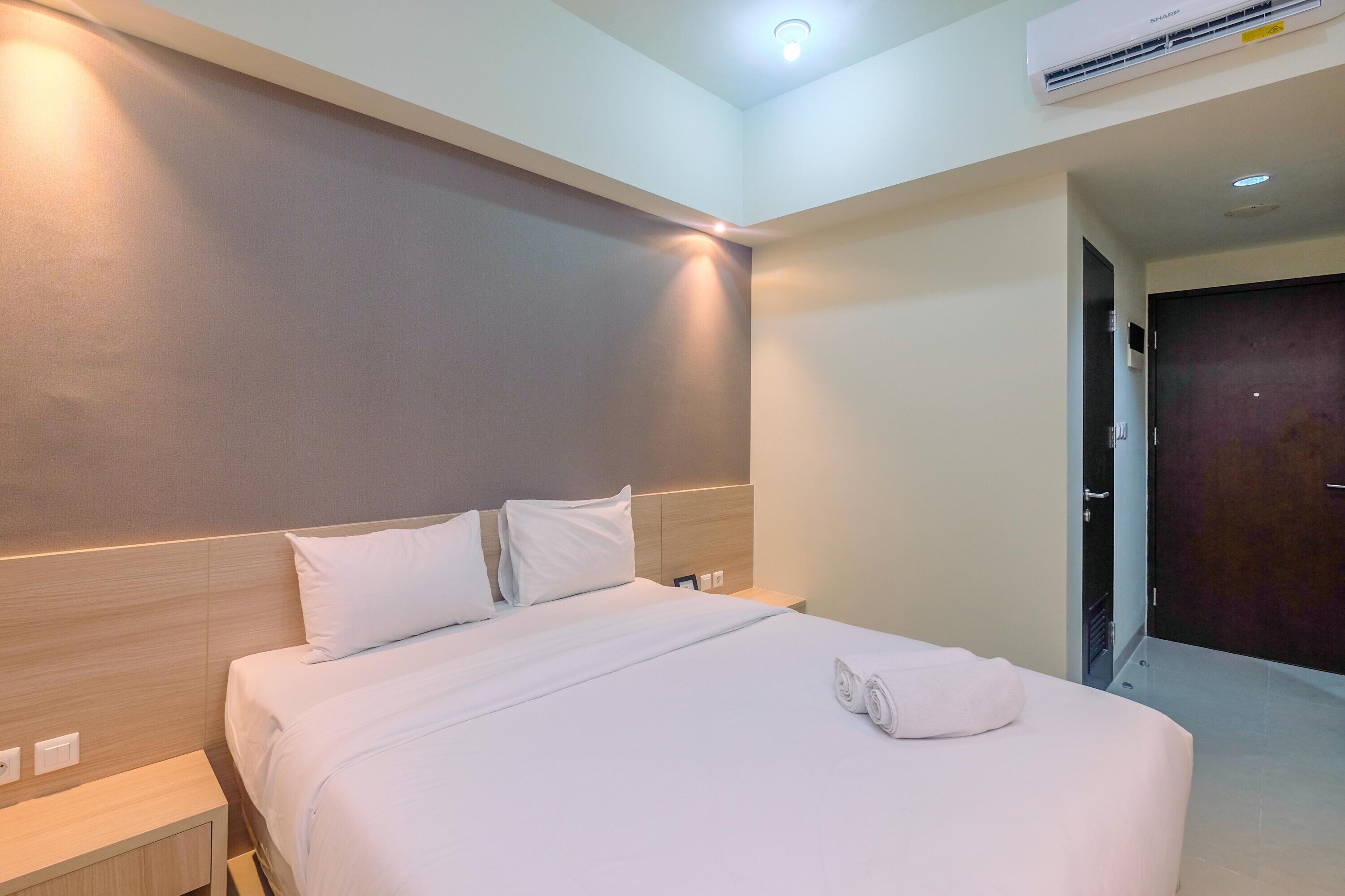 A neatly made bed with white linens and a rolled towel, a wooden headboard, a bedside table, and a wall-mounted air conditioner.