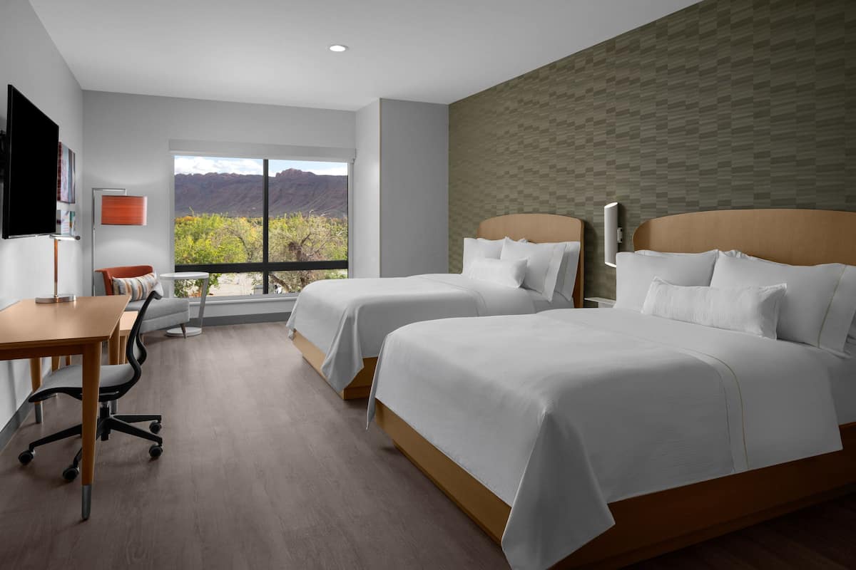 A hotel room with two beds, a desk, and a chair. There is a large window offering a view of mountains.
