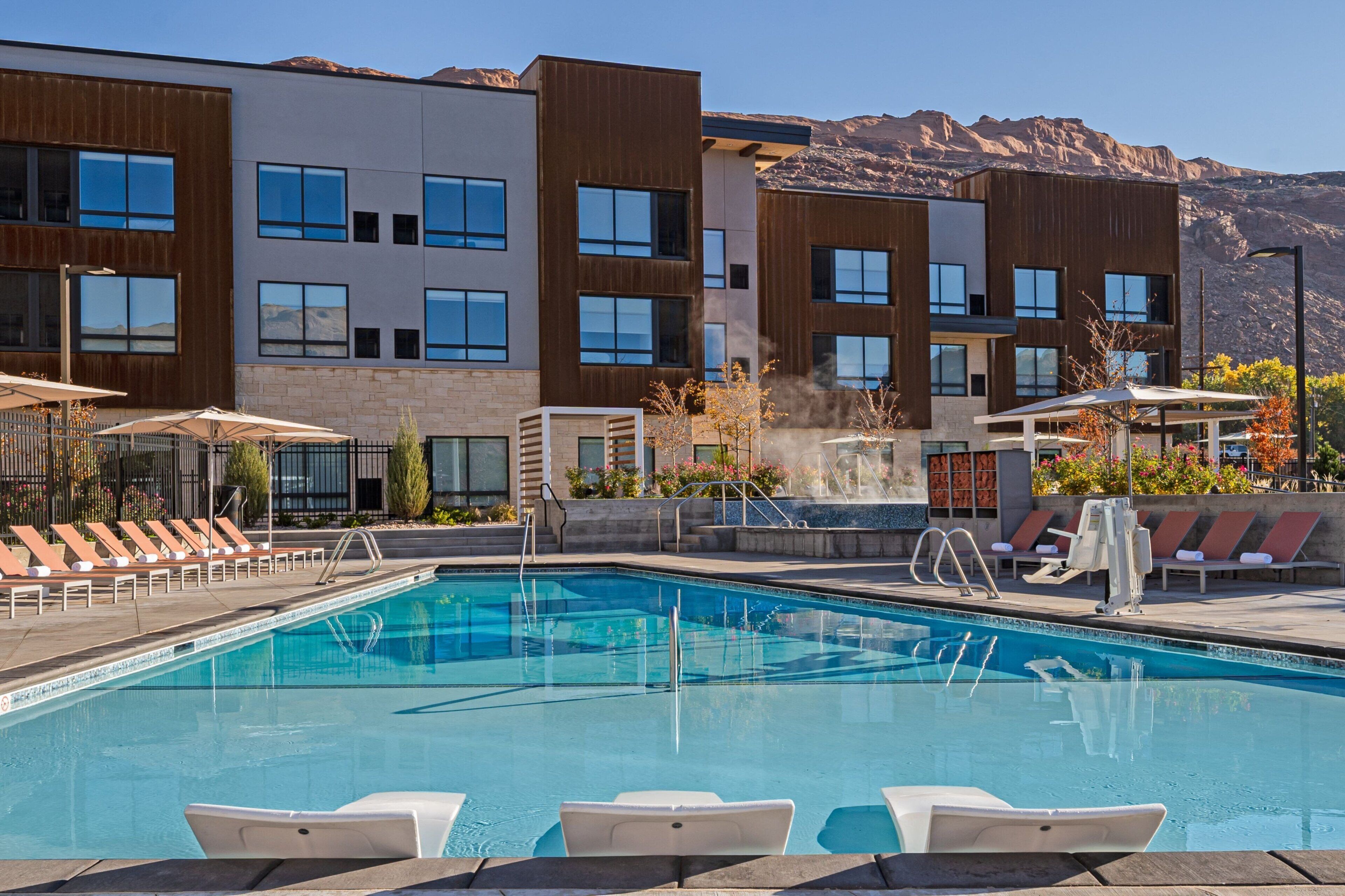 A modern hotel with a swimming pool, lounge chairs, and a mountain backdrop.